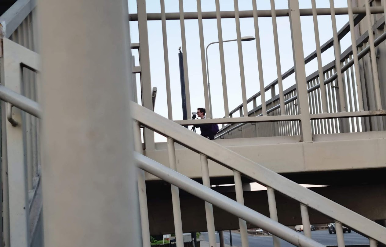 An image of a network of grey railings leading to an overpass. In the distance through a gap in the railings is artist Aislinn Evans capturing film through a camera.