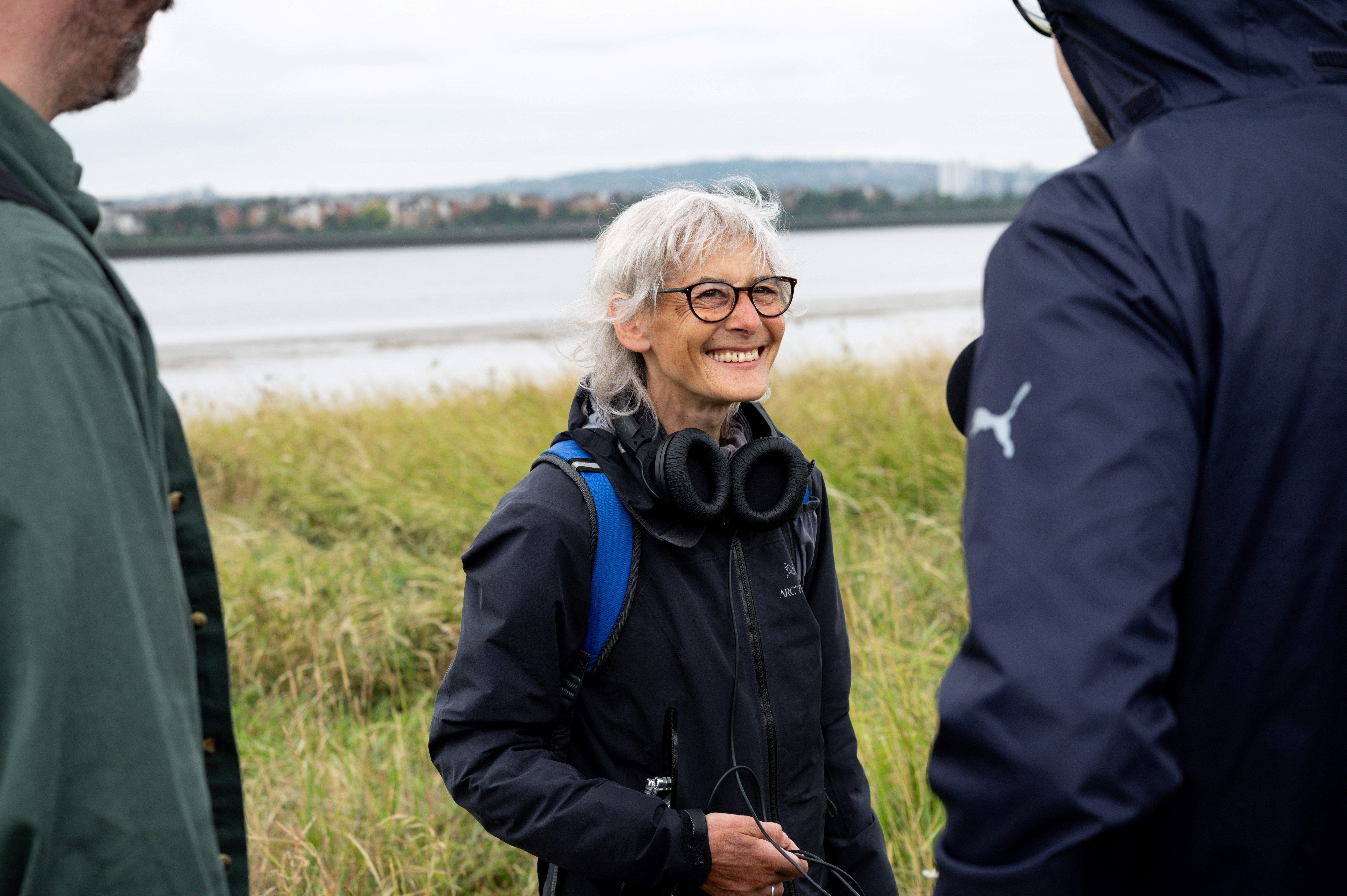 An image of artist Catherine Yass- a woman with white hair and black framed glasses, with a pair of headphones around her neck, in a blue rain mac with river behind her.